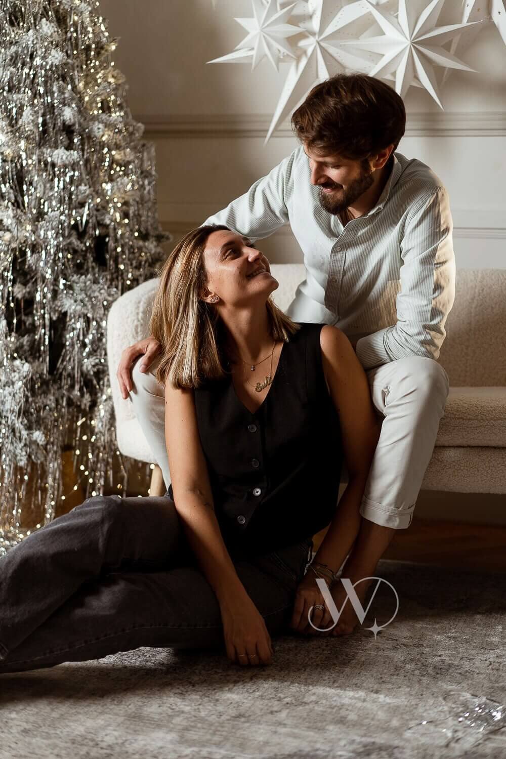 Pareja joven sentada en el suelo frente a un árbol de Navidad, abrazándose.