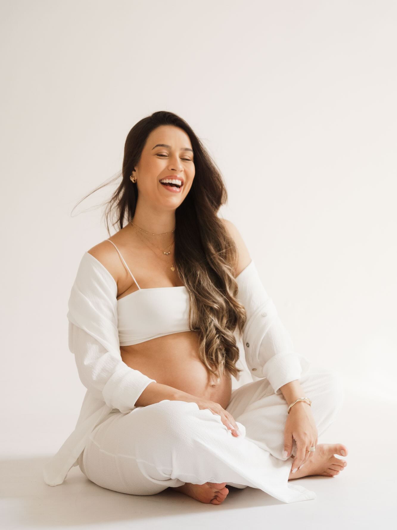 Embarazada riendo sentada en el suelo del estudio, fotos de embarazo con inseguridades superadas, look blanco
