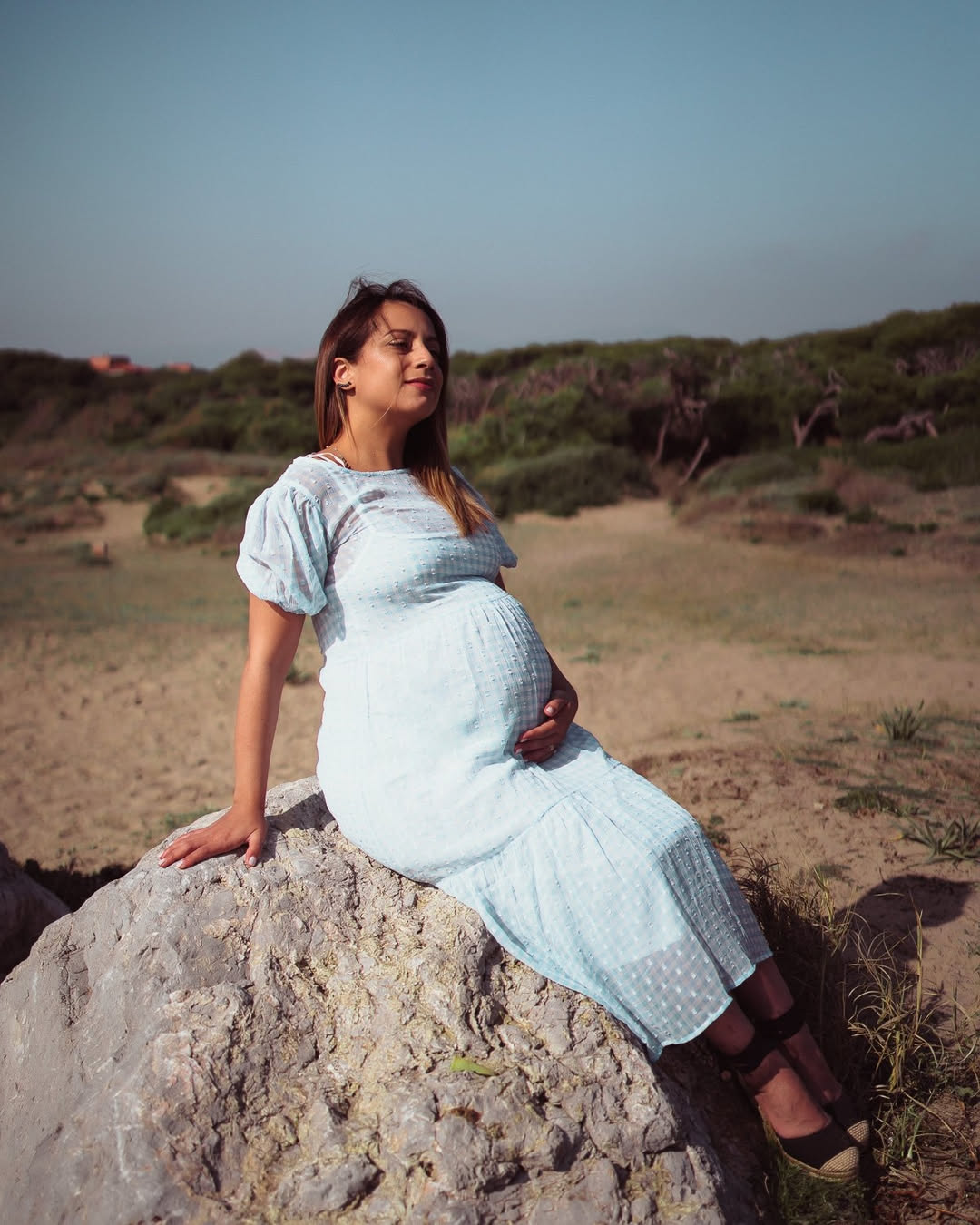 Pregnant woman sitting on a rock in a dune area, light blue dress