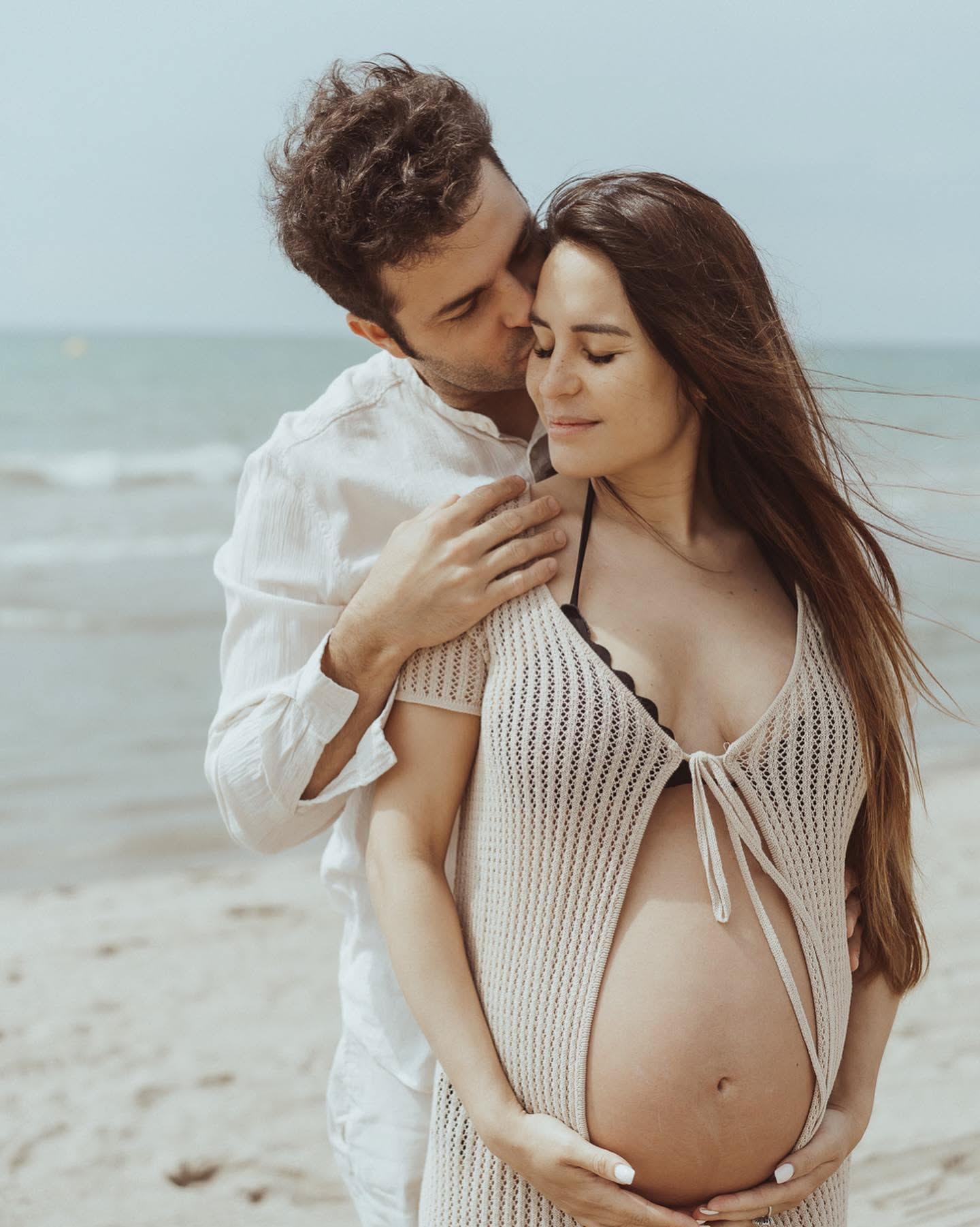 Pregnant woman with her partner at the beach, wind in their hair, maternity session Barcelona