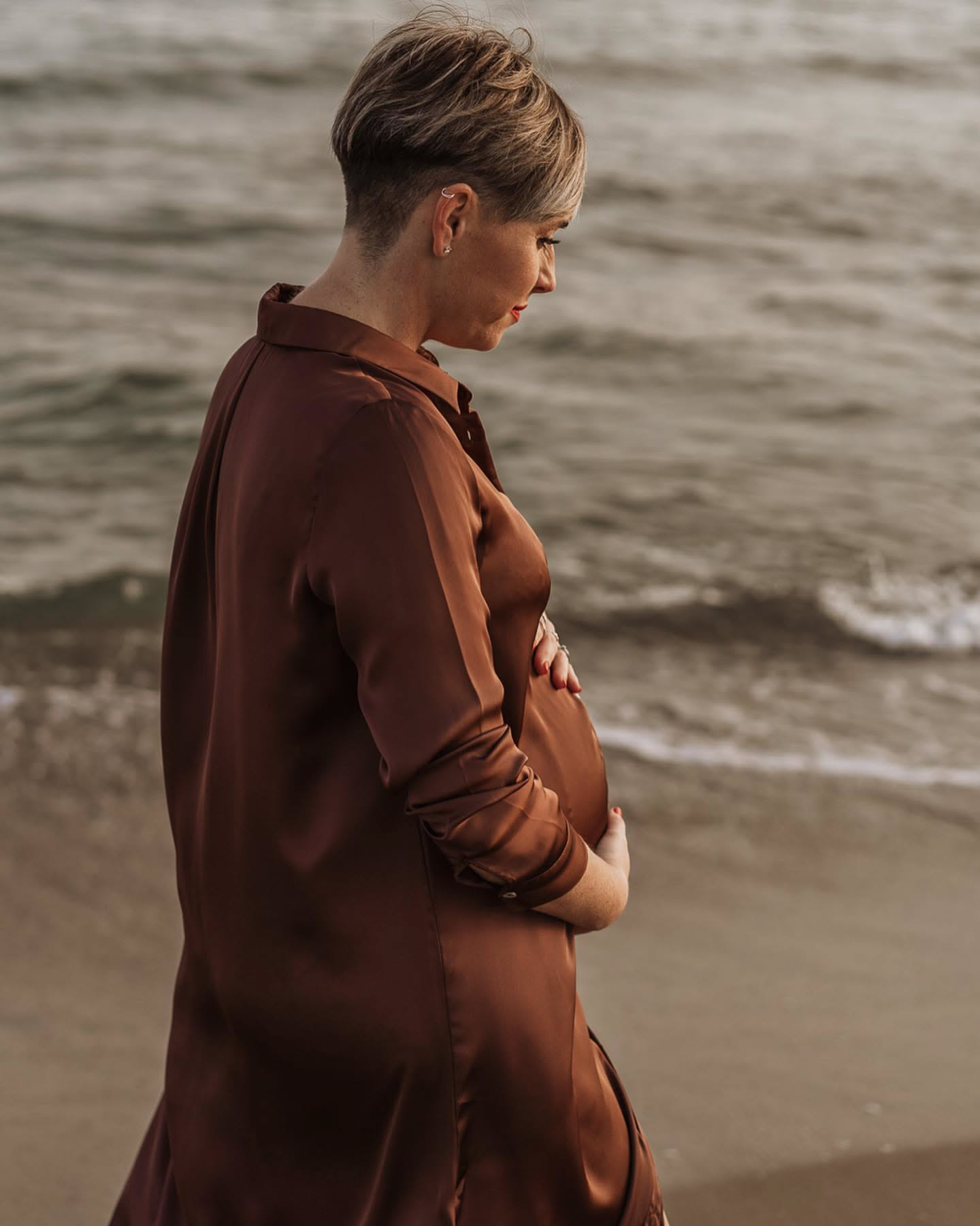 Pregnant woman in a brown dress at the beach at sunset, warm light on the sea