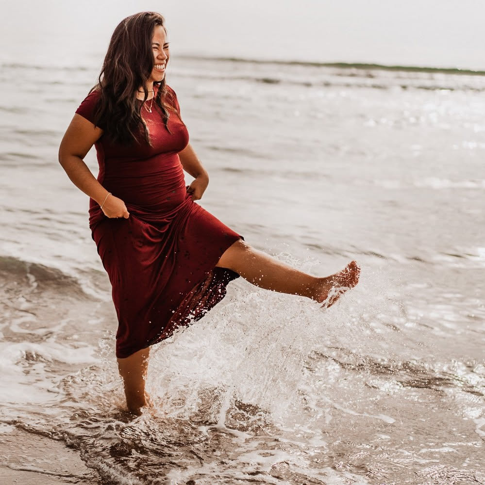 Pregnant woman laughing and playing in the waves, red dress at the shoreline