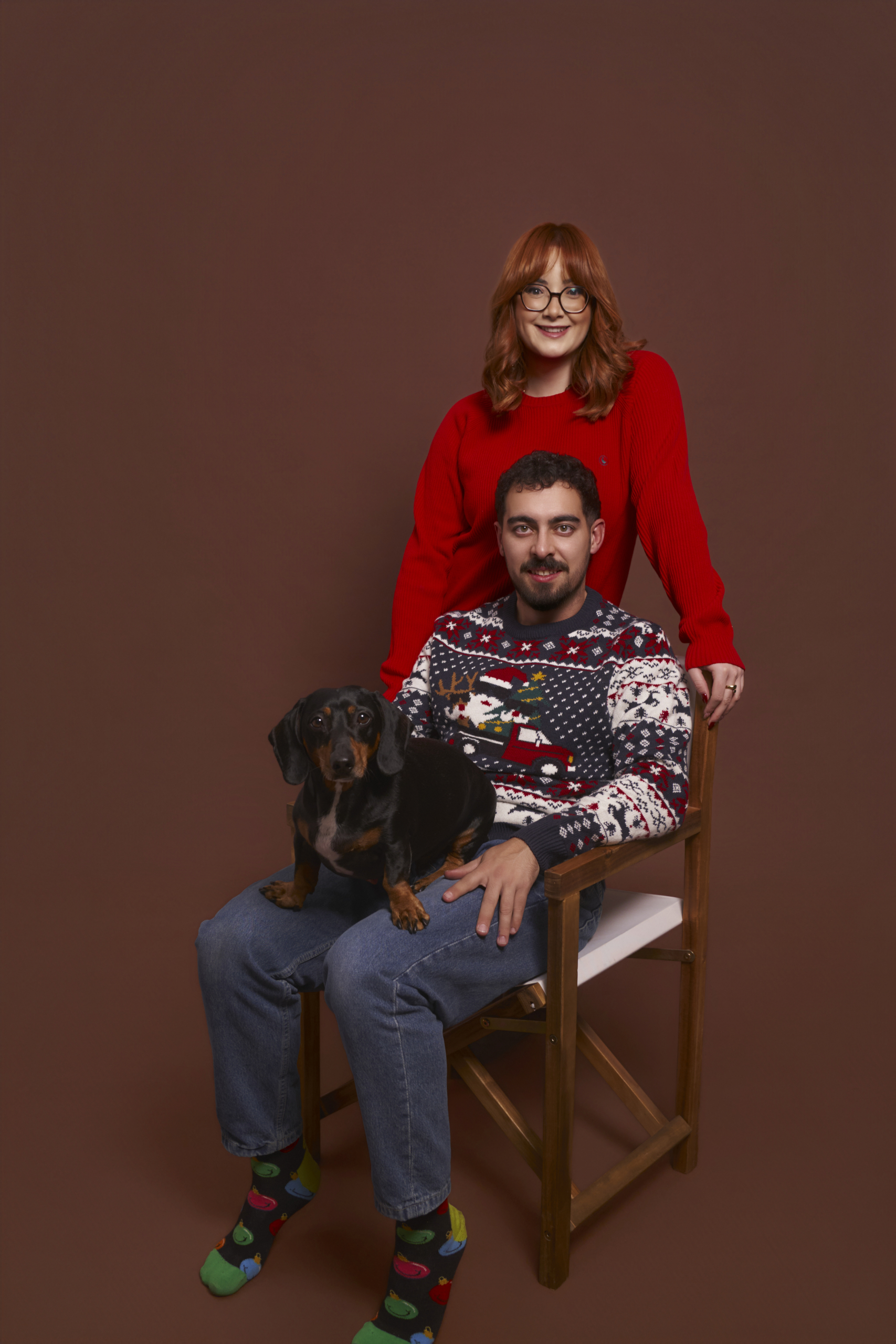 Pareja con jerseis navidenos y su perro salchicha sobre fondo rojo calido