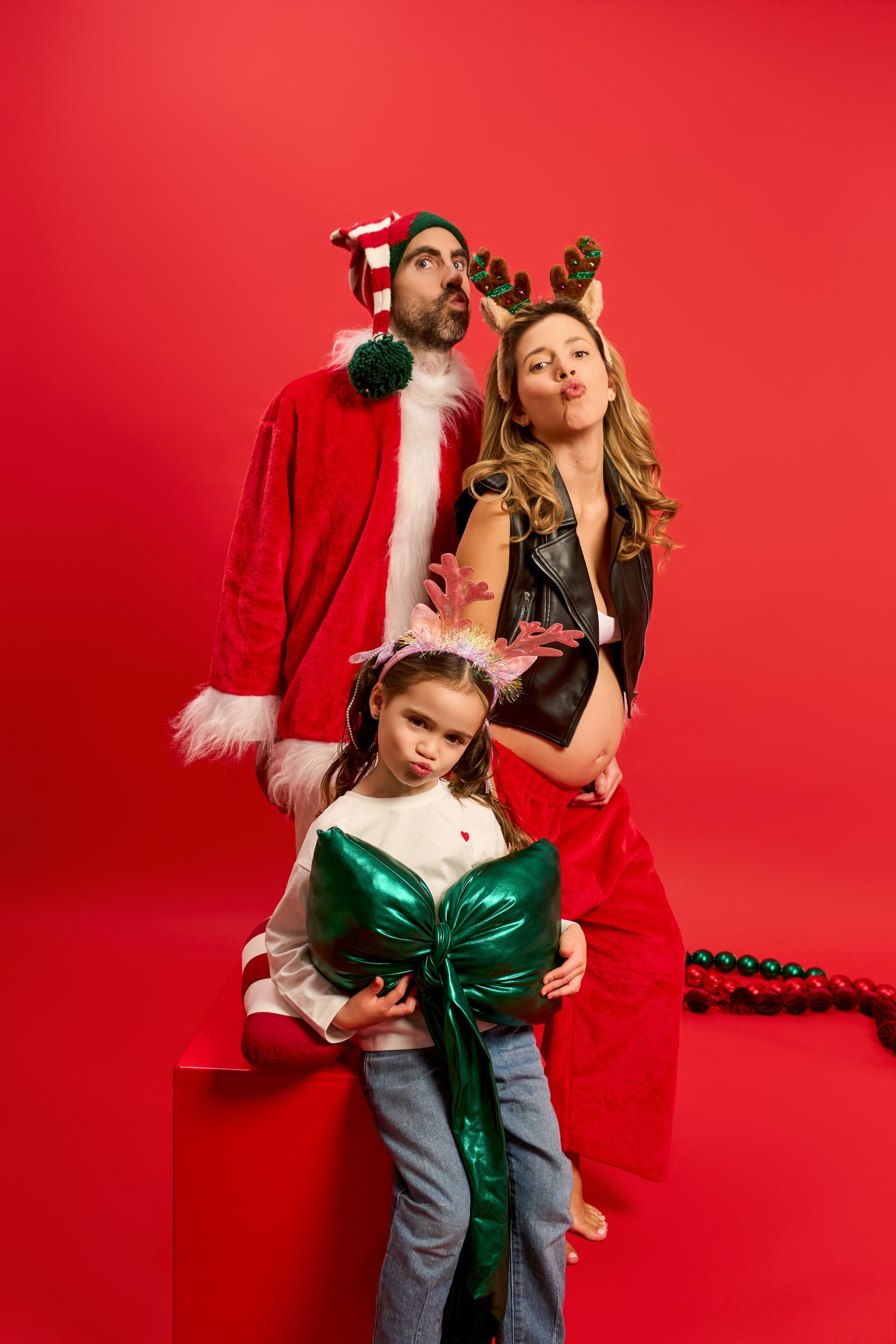 Pare amb jaqueta de Pare Noel, mare embarassada i nena amb banyes de ren i un llaç verd a l'estudi YOULO, fons vermell